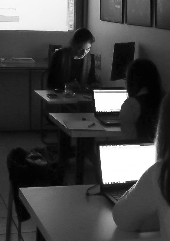 Melis Baloglu'nun karanlik bir ortamda aydinlik projeksiyon perdesi onunde basi hafif onundeki bilgisayara egik olarak silueti seciliyor, acik bilgisayar ekranlari onunde oturan iki kisi arkadan goruluyor